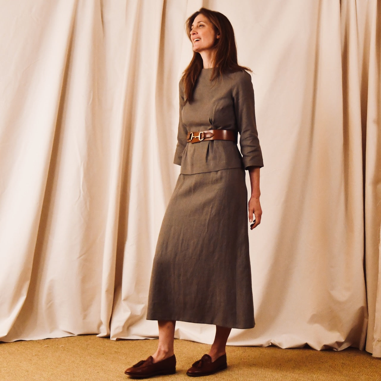 A woman stands in front of beige curtains wearing a tailored, gray, long-sleeved, ankle-length dress with a brown belt and loafers. She has straight, shoulder-length hair and is smiling while looking to the side.