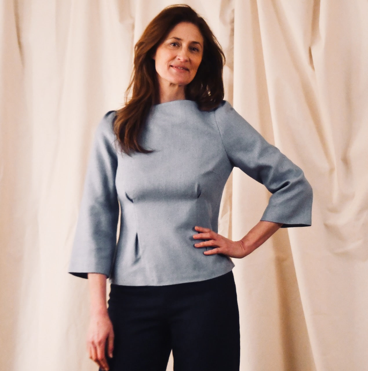 A woman with long brown hair stands in front of beige drapes, wearing a British-made, light blue long-sleeved top with a fitted waist and black pants. One hand is on her hip as she smiles softly at the camera.