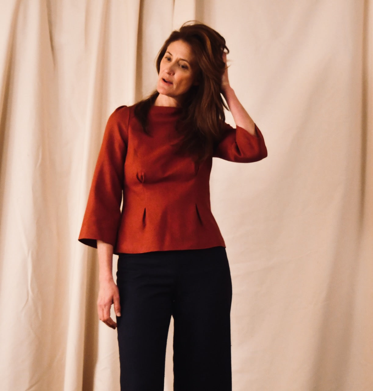 A woman with long brown hair wearing a tailored red top and black pants stands in front of a beige curtain, touching her hair with one hand and looking slightly to the side.