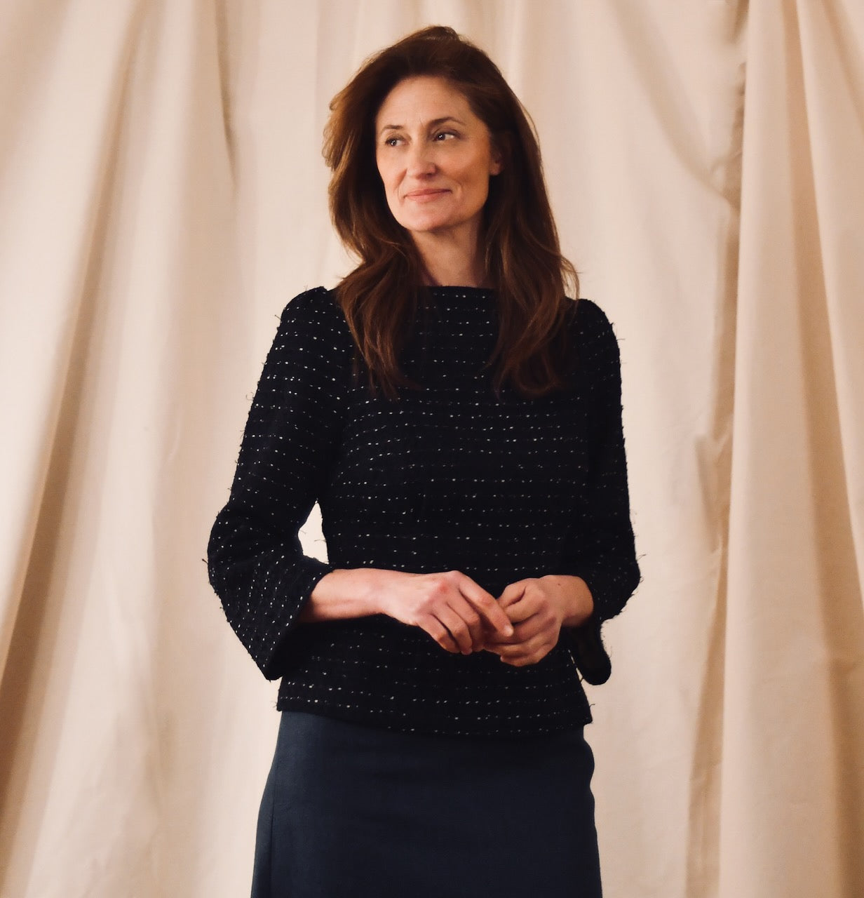 A woman with long brown hair stands in front of beige curtains, wearing a British-made black textured long-sleeve top and a dark skirt, smiling softly while looking slightly to the side.