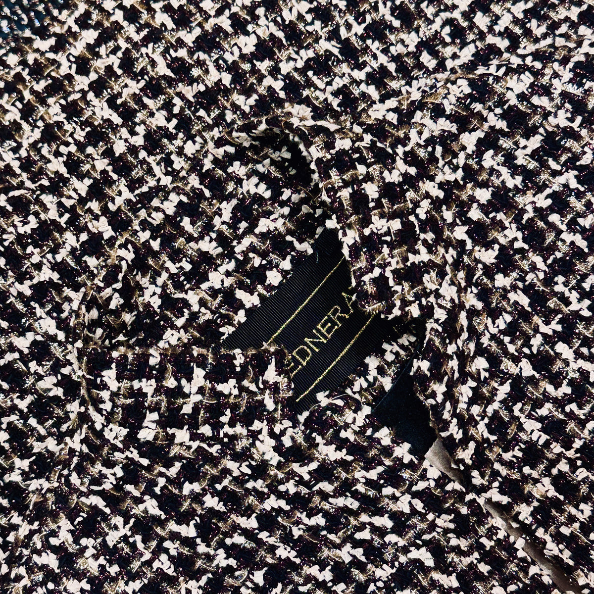 A close-up of a tailored black, white, and metallic tweed fabric jacket with a visible garment label partially showing gold text.