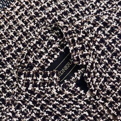 A close-up of a tailored black, white, and metallic tweed fabric jacket with a visible garment label partially showing gold text.