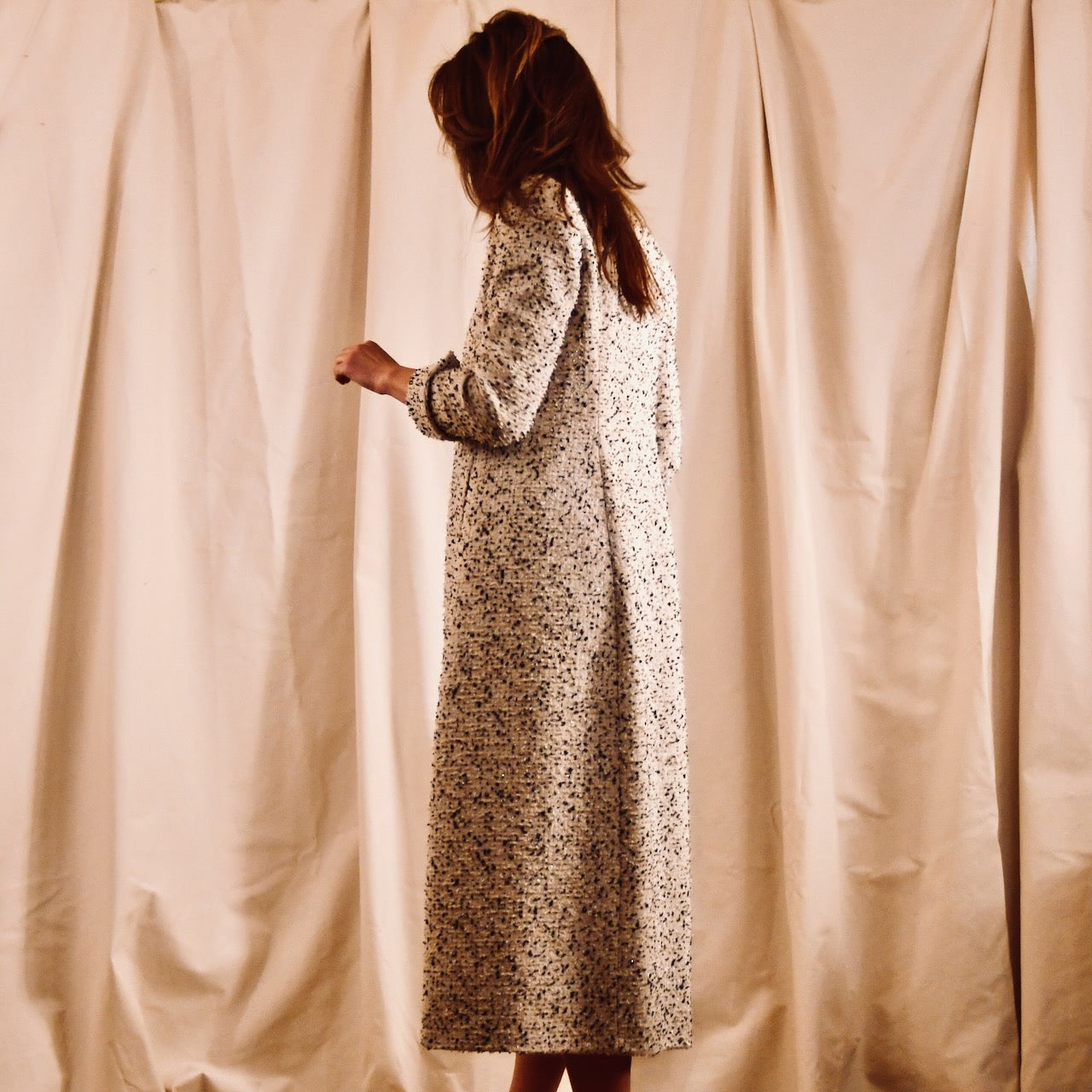 A woman in a long, hand-made, light-colored patterned dress stands sideways with her head slightly turned down. She is in front of a beige, wrinkled fabric backdrop.