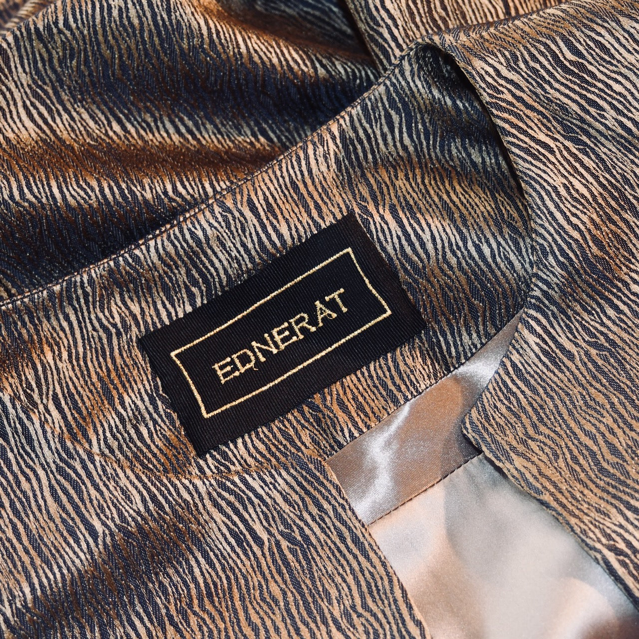 A close-up of a hand-made EDNERAT garment label sewn onto fabric with a textured, wavy blue and gold pattern. The silvery gray inside lining is visible.