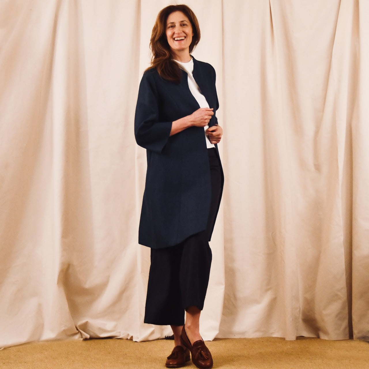 A woman with long brown hair smiles in front of beige curtains, wearing the EDNERAT Trapeze Swing Coat in Navy Heavier Weight Linen over a white top, black wide-leg pants, and brown loafers on a tan carpet.