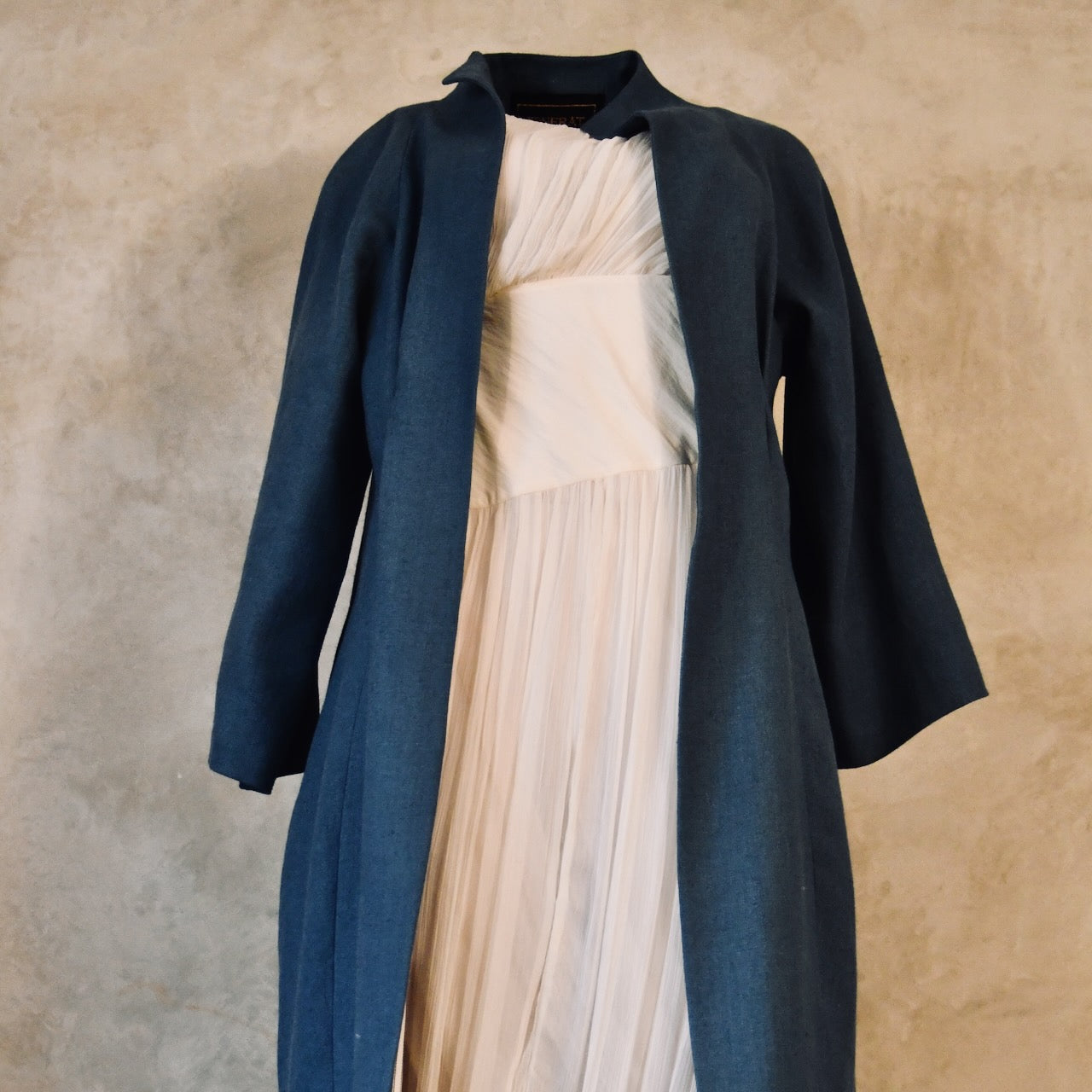 A mannequin dressed in a flowing, pleated white dress layered under a long, tailored dark blue open coat, set against a neutral textured background.