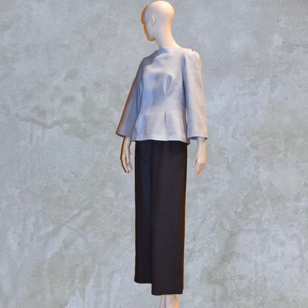 A Tailored Boat Neck Blouse in Pale Blue Silk Matka with contrasting trousers, posed sideways. Handmade in Britain. A refined ensemble defined by elegant lines.