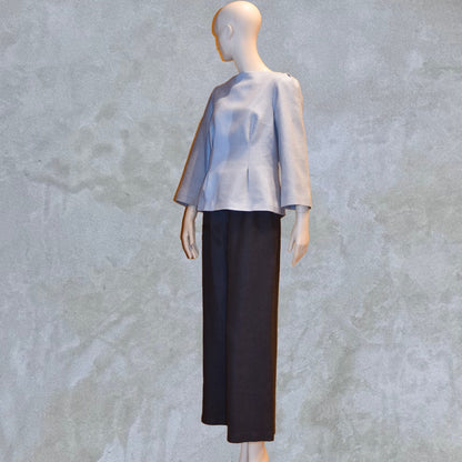 A Tailored Boat Neck Blouse in Pale Blue Silk Matka with contrasting trousers, posed sideways. Handmade in Britain. A refined ensemble defined by elegant lines.
