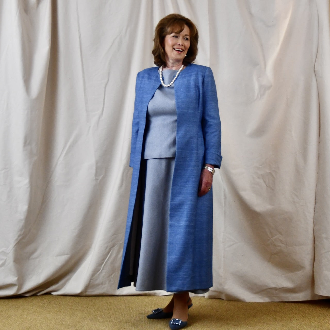 Tailored, Fitted Hourglass shaped coat in a summer-weight Silk Madras in Blue. This is a long-sleeved, calf-length blue summer coat with an hourglass shape. Shown from the side-front. A perfect summer coat for weddings and special occasions. Handmade in Britain.