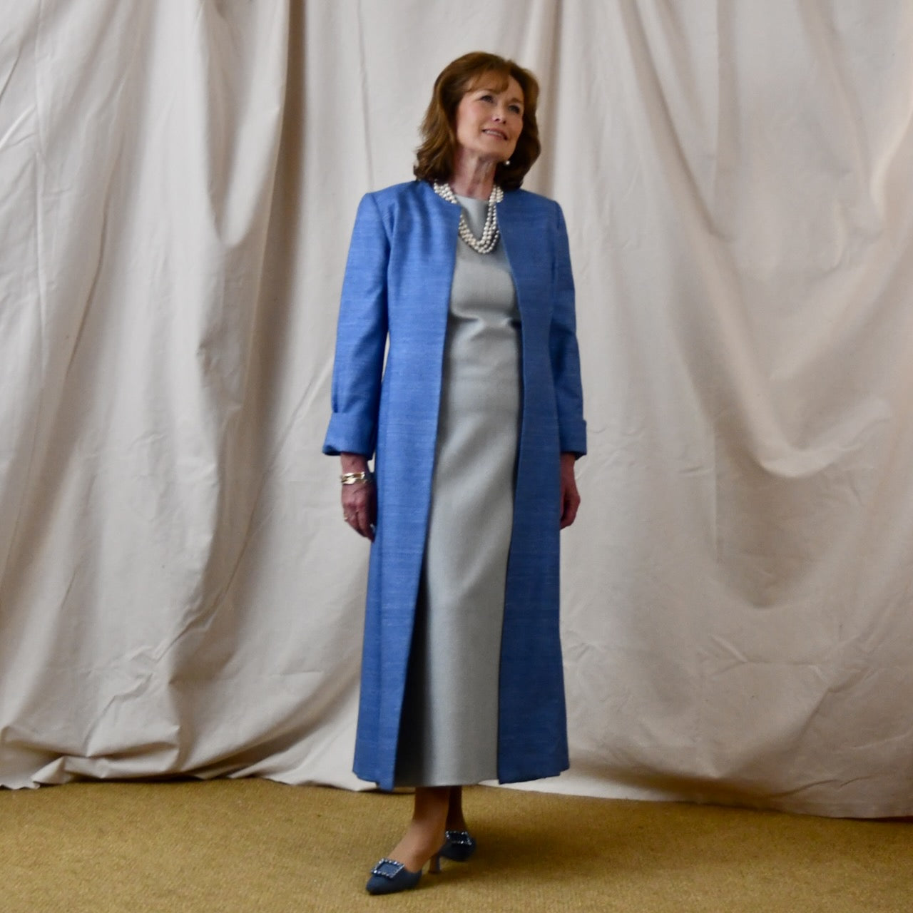 Tailored, Fitted Hourglass shaped coat in a summer-weight Silk Madras in Blue. This is a long-sleeved, calf-length blue summer coat with an hourglass shape. Shown from the side-front. A perfect summer coat for weddings and special occasions. Handmade in Britain.