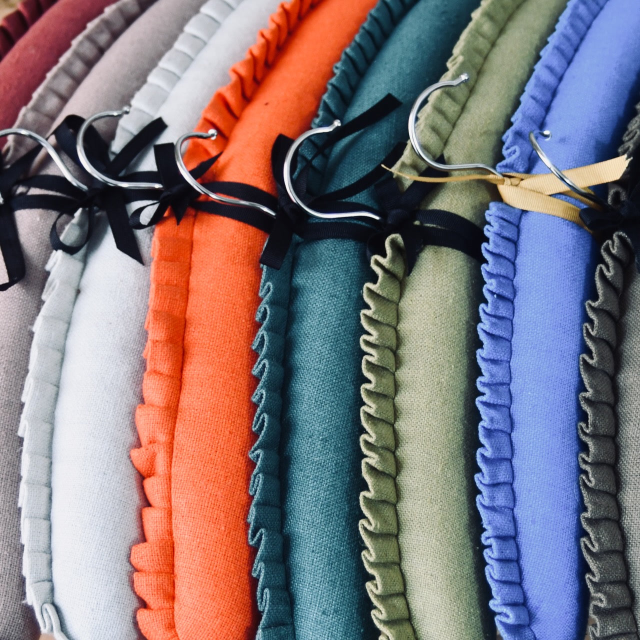 A close-up of hand-made padded clothes hangers in various colors—beige, red, orange, green, and blue—each with a ruffled edge and tied with a small ribbon near the hook.