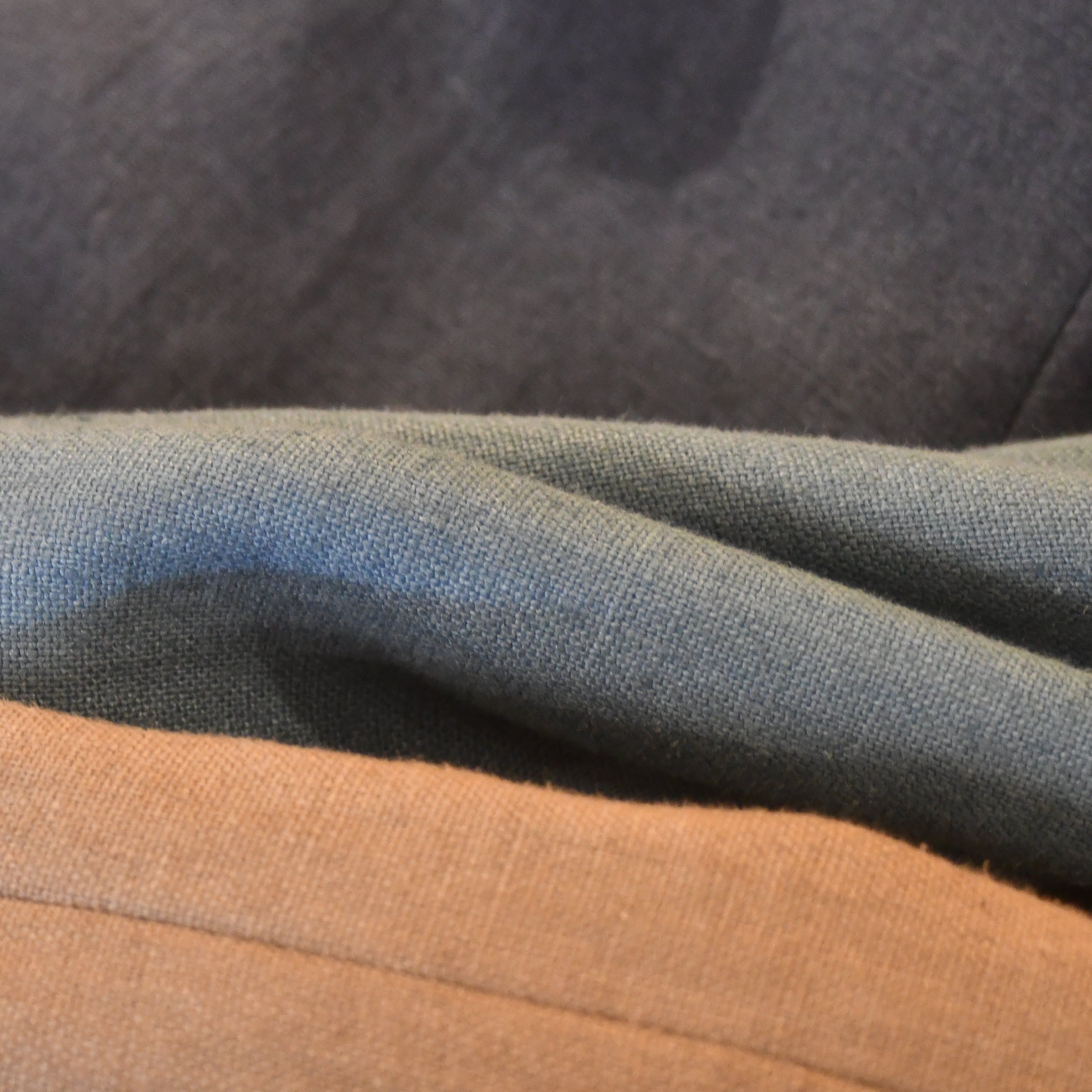 Three pieces of hand-made fabric in dark gray, muted green, and light brown are layered and draped, showcasing their textures and natural fibers from EDNERAT.