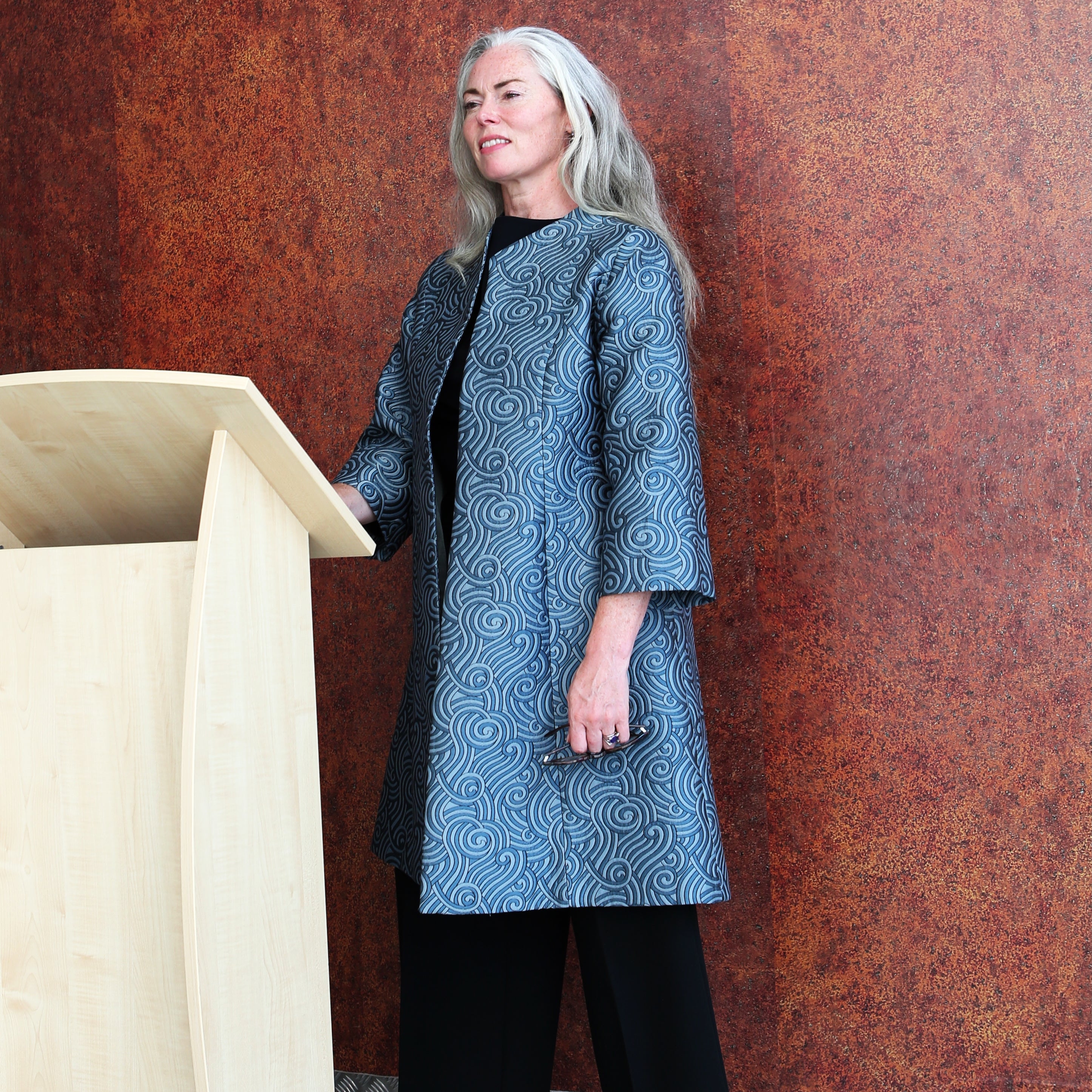 A woman with long gray hair stands at a light wooden podium, wearing a tailored, hand-made gray coat over black clothing, against a textured brown wall.