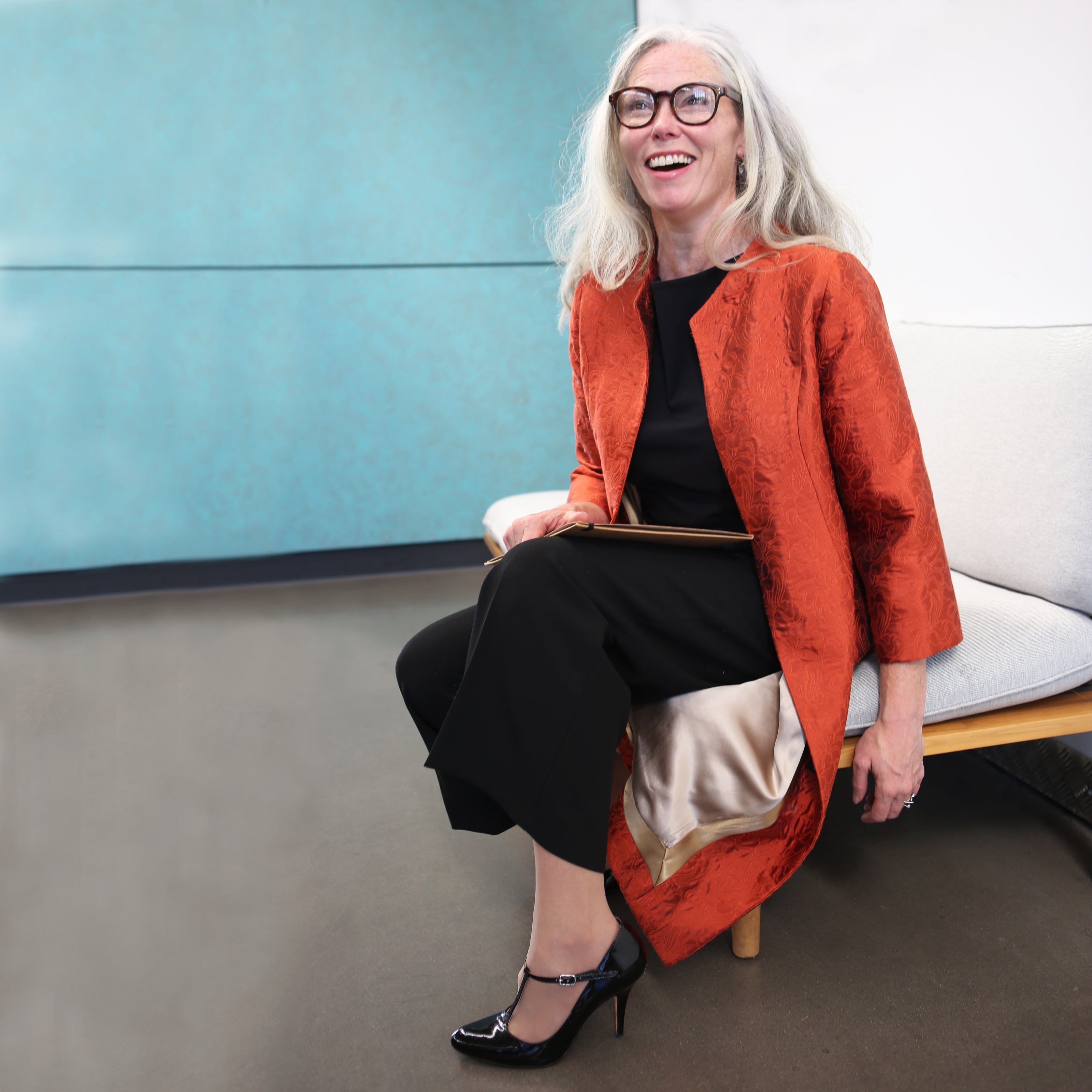 Smiling older woman with long gray hair and glasses sits on a modern bench, wearing a tailored black outfit with a shiny orange coat and black heels, in a bright room with a blue wall.