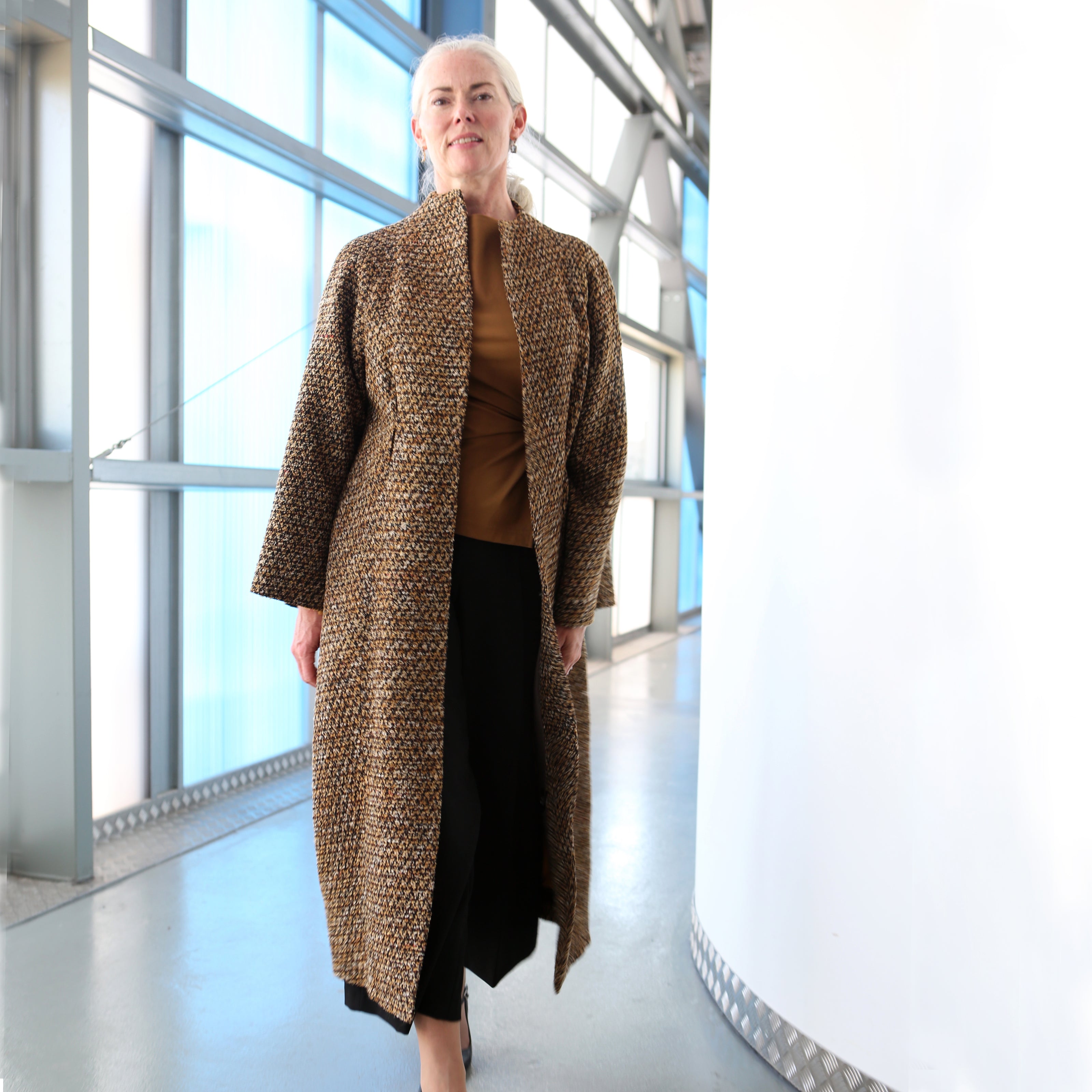 A woman with gray hair pulled back stands indoors, smiling in a modern, sunlit hallway with large windows. She wears a tailored, hand-made patterned coat over a brown top and black pants.