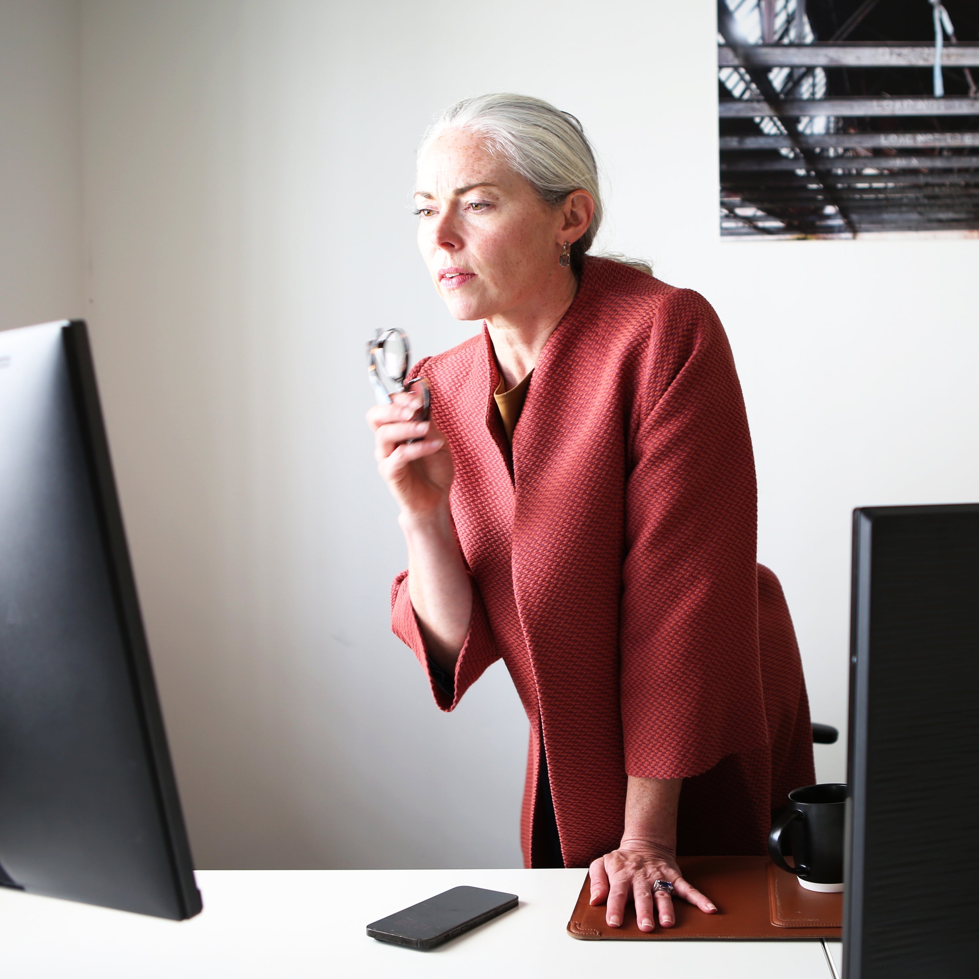 An older woman with gray hair in a ponytail stands at an office desk, holding glasses and looking intently at a computer monitor. A phone, a mug, and another monitor are on the tailored workspace.