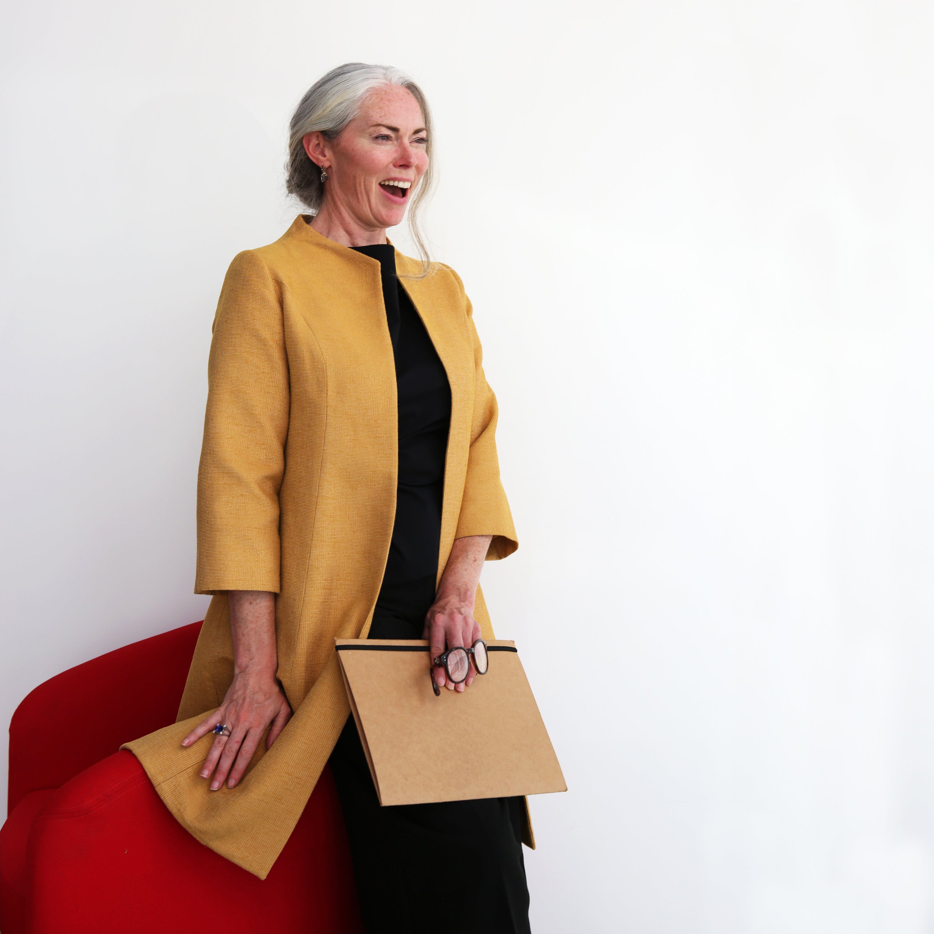 A smiling older woman with long gray hair stands beside a red chair, wearing a mustard yellow coat over a black dress. She holds a closed laptop and glasses, showcasing her Bespoke style. The background is plain white.