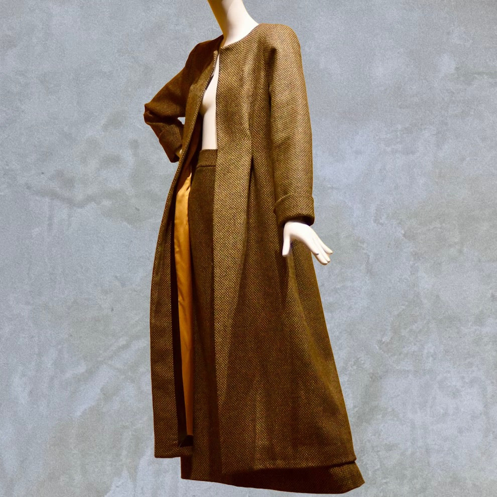 Longer length Skirted Hourglass Coat in Orange-Green Tweed. This shaped coat uses clever tailoring details to create a wonderfully flattering and easy to way skirted shape. With waist-enhancing tucks it will flatter the most testing of frames.  Entirely made in Great Britain. Worn open with matching skirt.