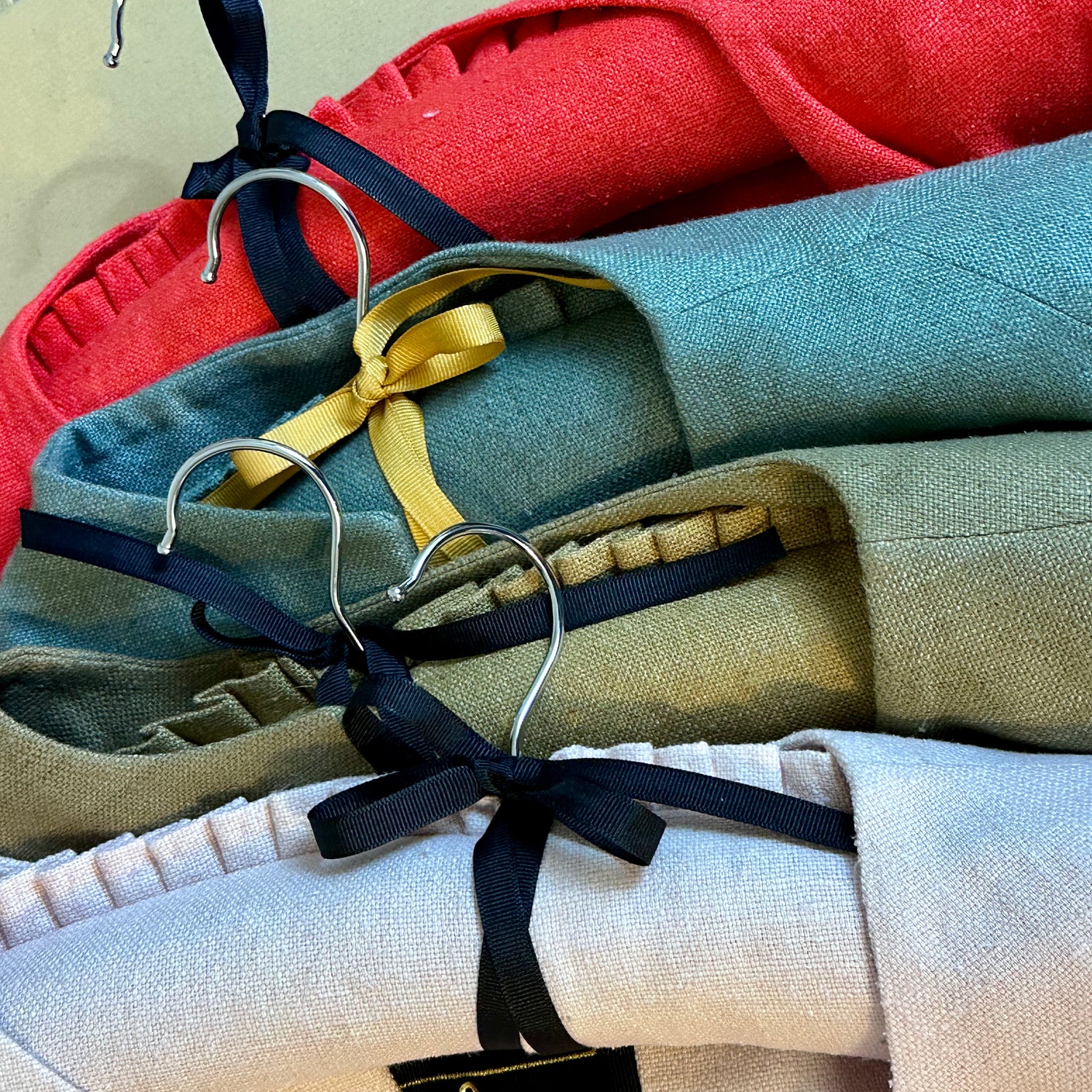 Four neatly folded, British-made linen garments in red, teal, olive green, and beige hang on metal hangers topped with black ribbon bows and a yellow ribbon, viewed from above.