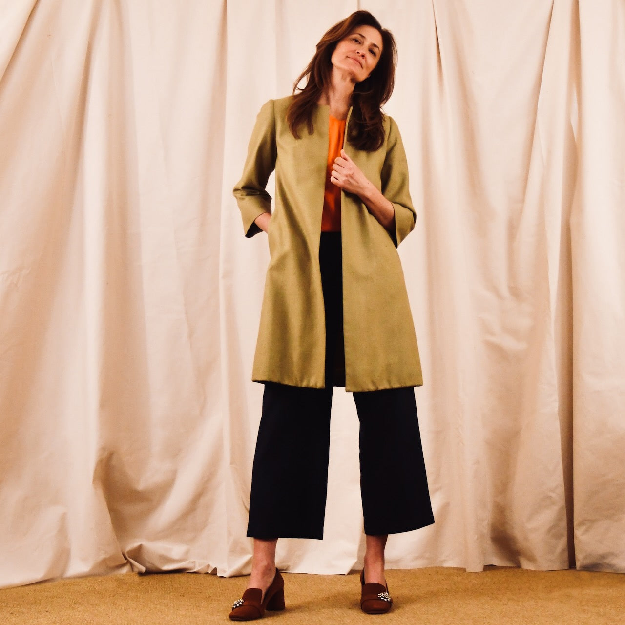  Trapeze Coat in Pea Green Silk Matka, paired with an orange top, black wide-leg pants, and brown shoes. Handmade in Britain.