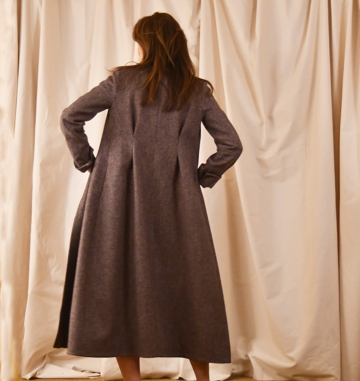 Long fitted tweed coat in pink colour. The ankle-length coat showcases a gray-pink textured tweed, hourglass tailoring, fitted seams, and back tucks. Handmade in Britain. Rear View.