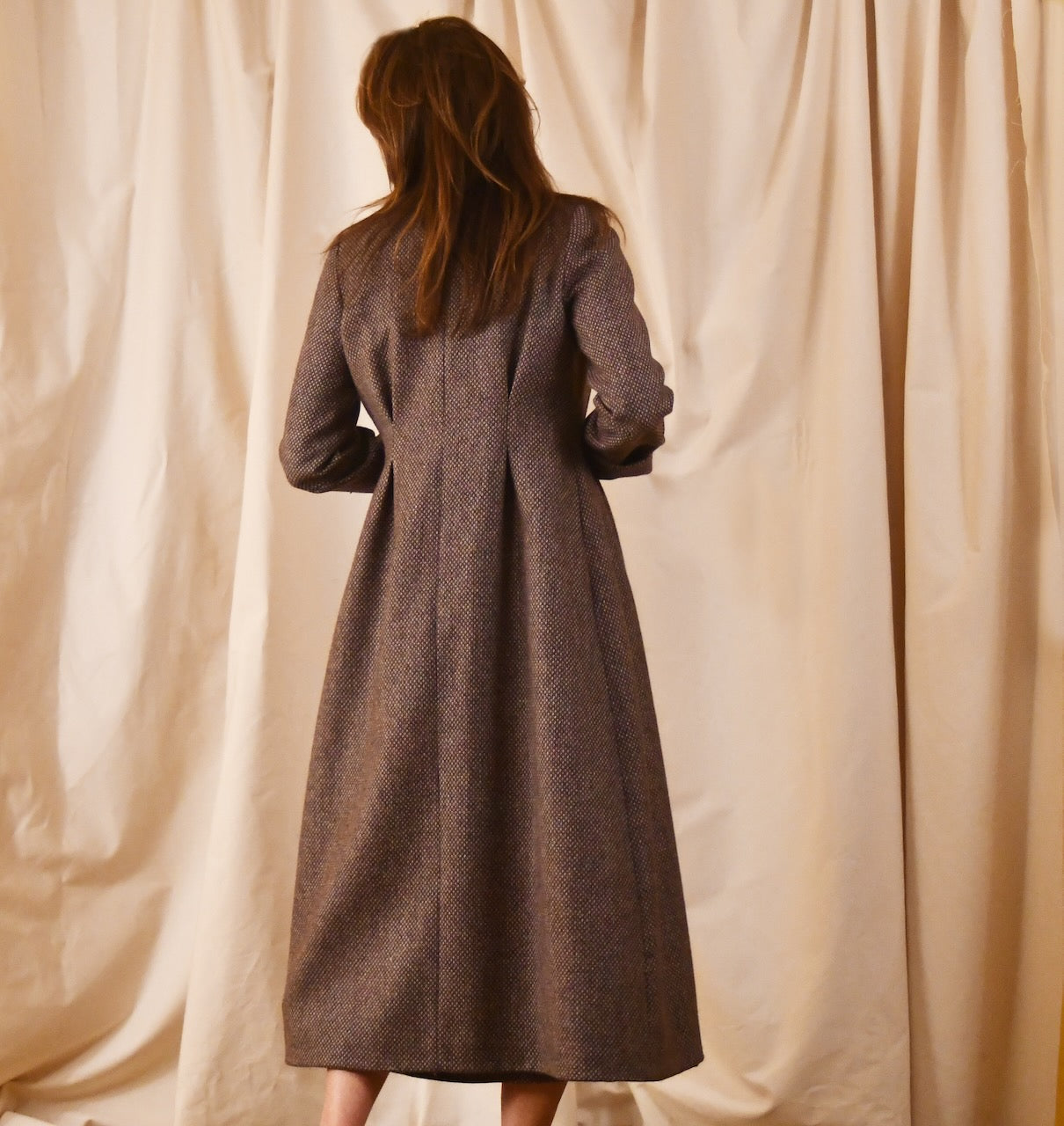 Long fitted tweed coat in pink colour. The ankle-length coat showcases a gray-pink textured tweed, hourglass tailoring, fitted seams, and back tucks. Handmade in Britain.
