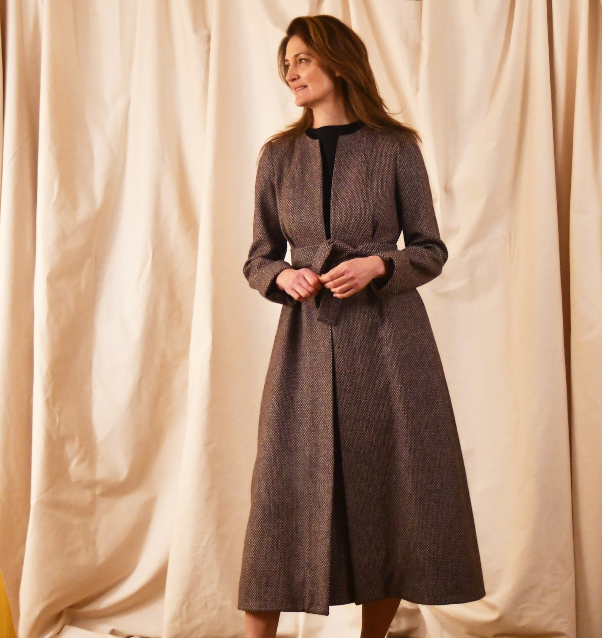 Long fitted tweed coat in pink colour. The ankle-length coat showcases a gray-pink textured tweed, hourglass tailoring, fitted seams, and back tucks. Handmade in Britain. Front View.
