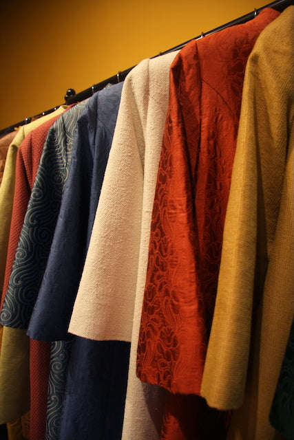 A row of colorful, textured hand-made garments on hangers—including red, white, blue, and yellow fabrics—displayed against a warm yellow background.