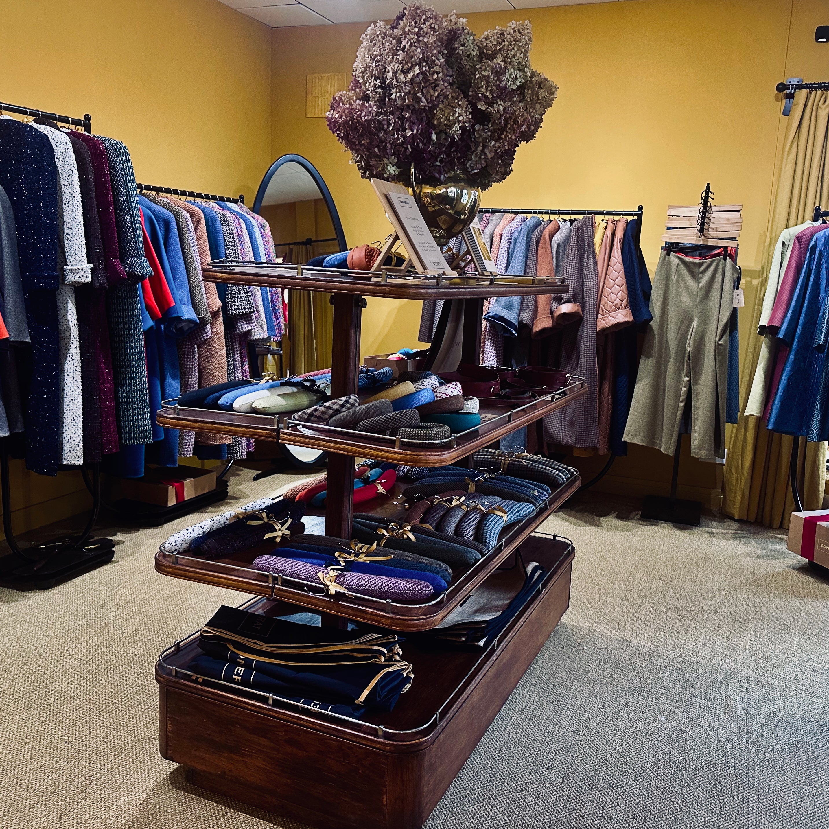 A boutique clothing store featuring racks of colorful clothes and a central wooden display with hand-made shirts, sweaters, ties, and accessories—topped with a vase of dried flowers.