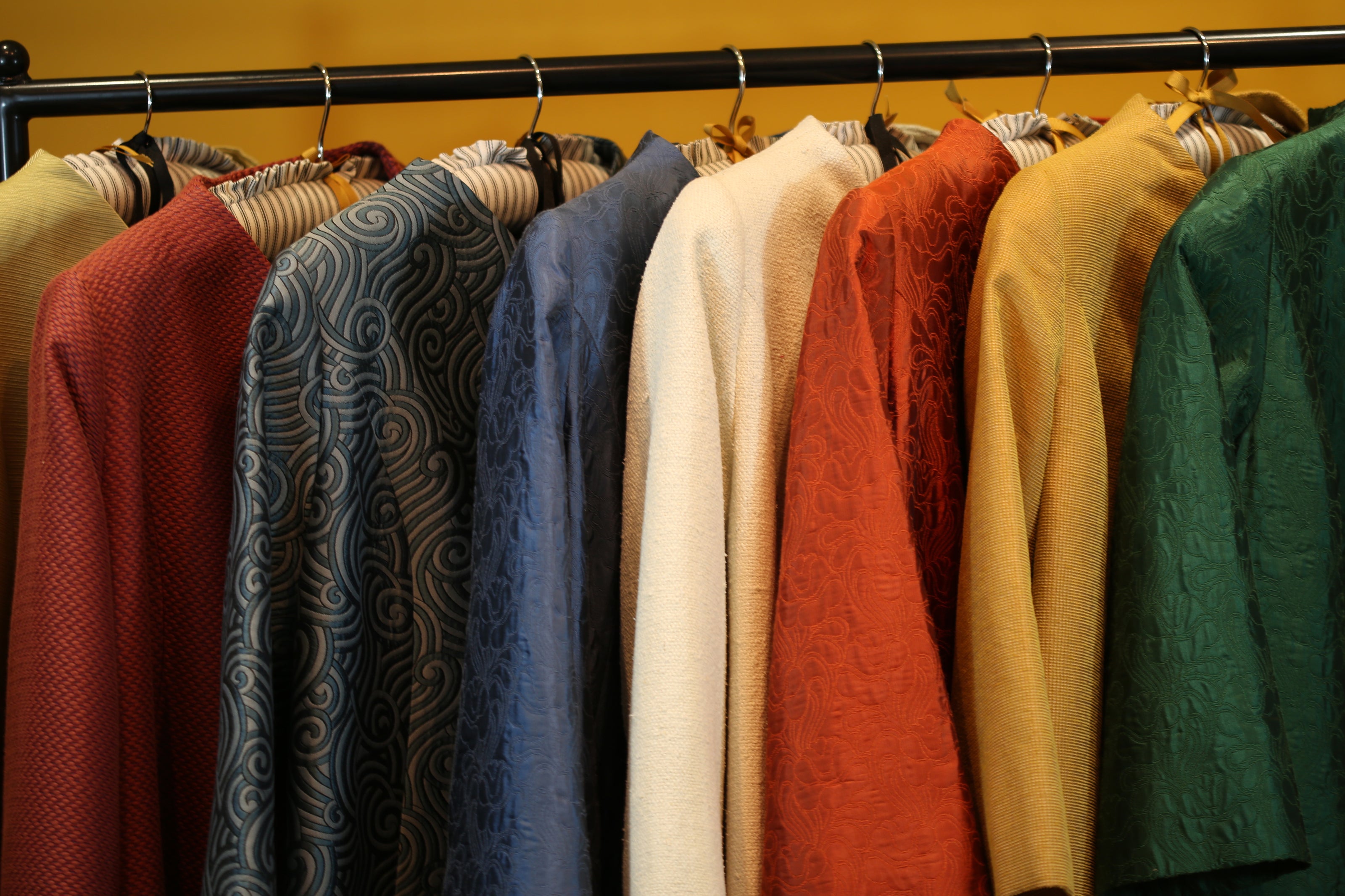 A row of colorful, hand-made jackets and shirts hang on wooden hangers from a black clothing rack, featuring patterns and solid colors including red, blue, white, orange, and yellow.
