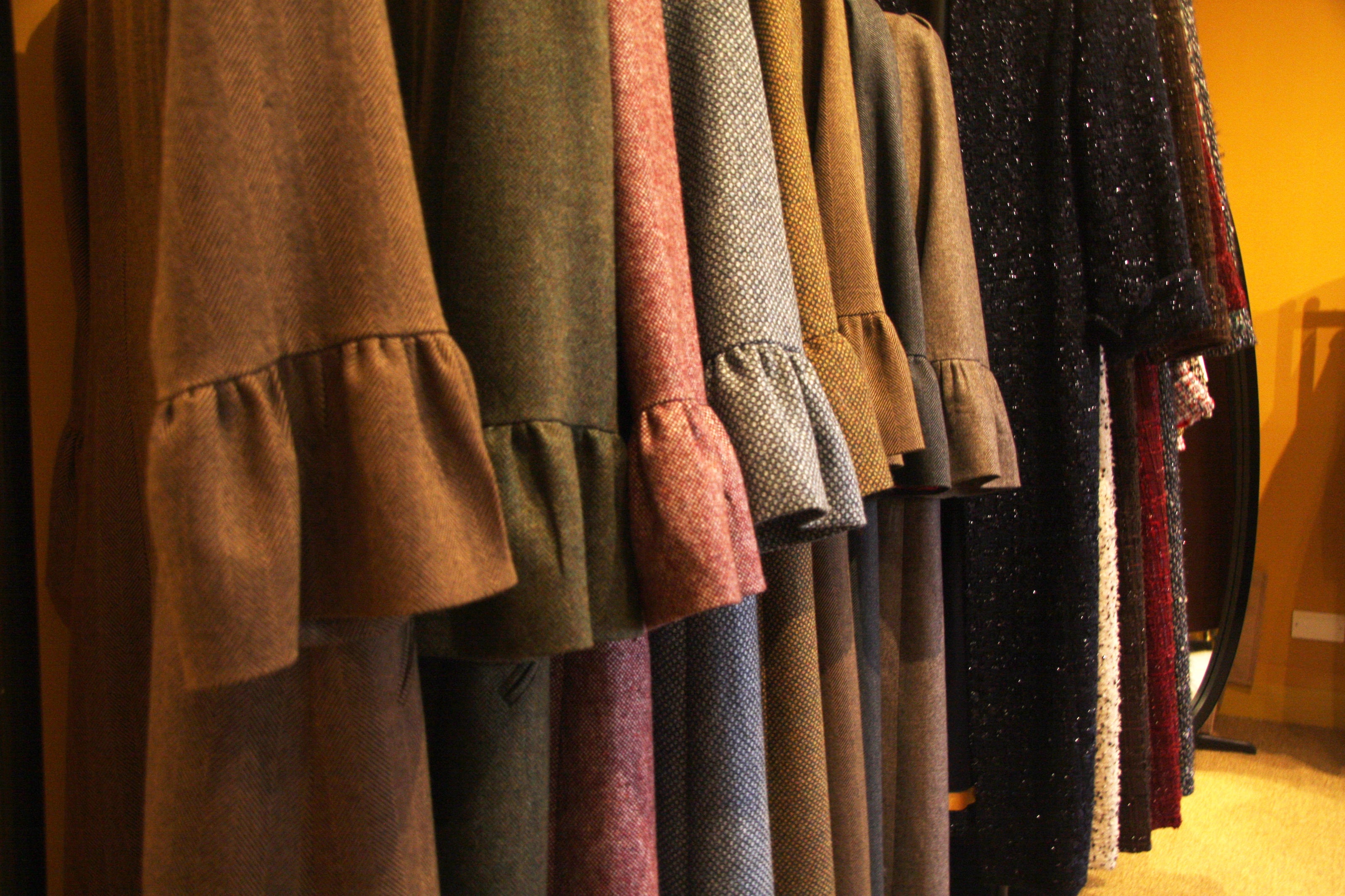 A row of bespoke, long-sleeved ruffled dresses in various earthy colors and textures hangs on a clothing rack against a warm yellow wall.