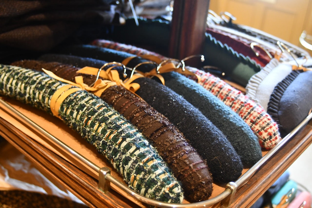 A row of hand-made padded clothes hangers covered in various textured and colorful fabrics, neatly arranged on a wooden shelf in a store or closet.