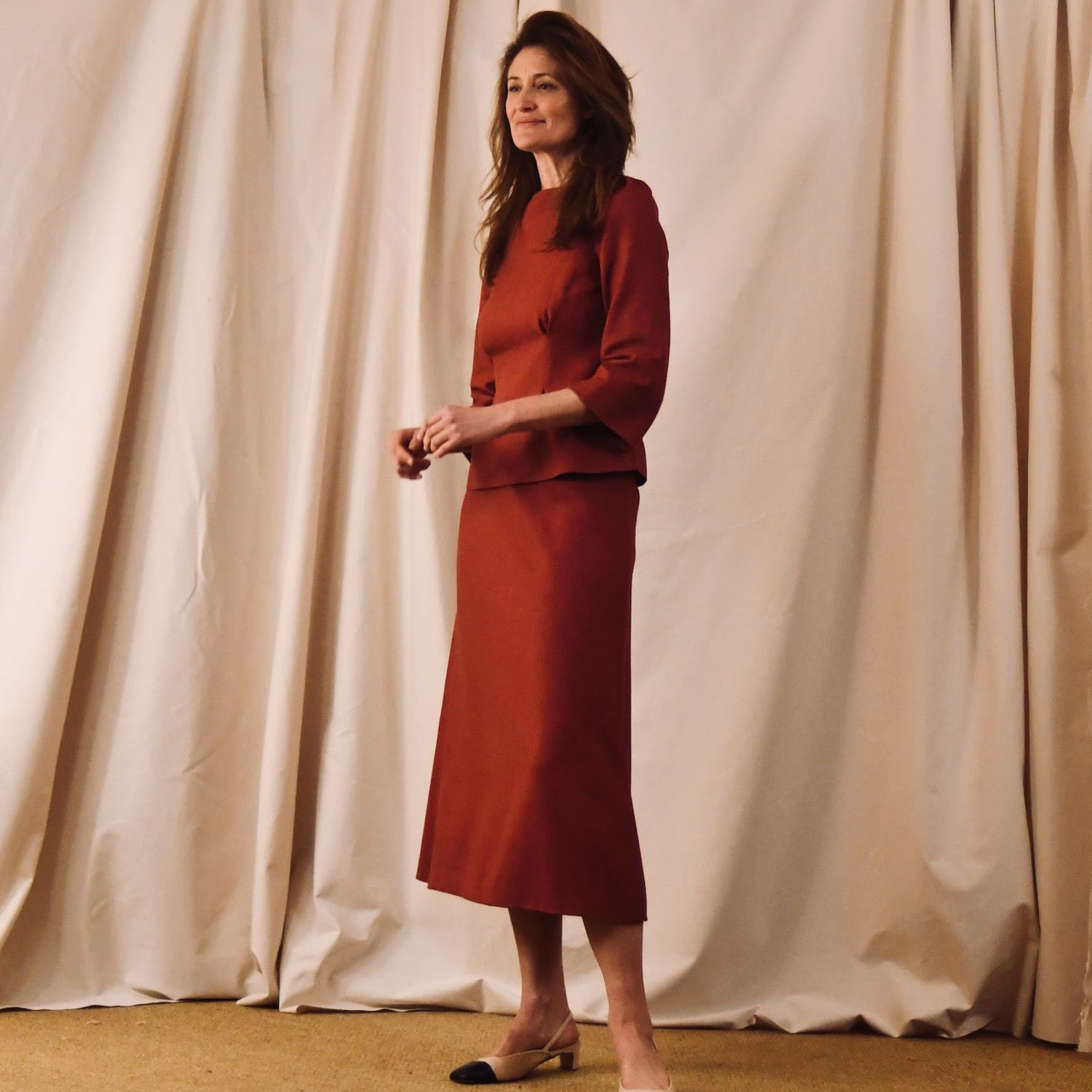 A woman stands in front of beige curtains, wearing a tailored, rust-colored long-sleeved top with a matching midi skirt from EDNERAT and beige and black low-heeled shoes. She smiles, her long wavy brown hair cascading over her shoulders.