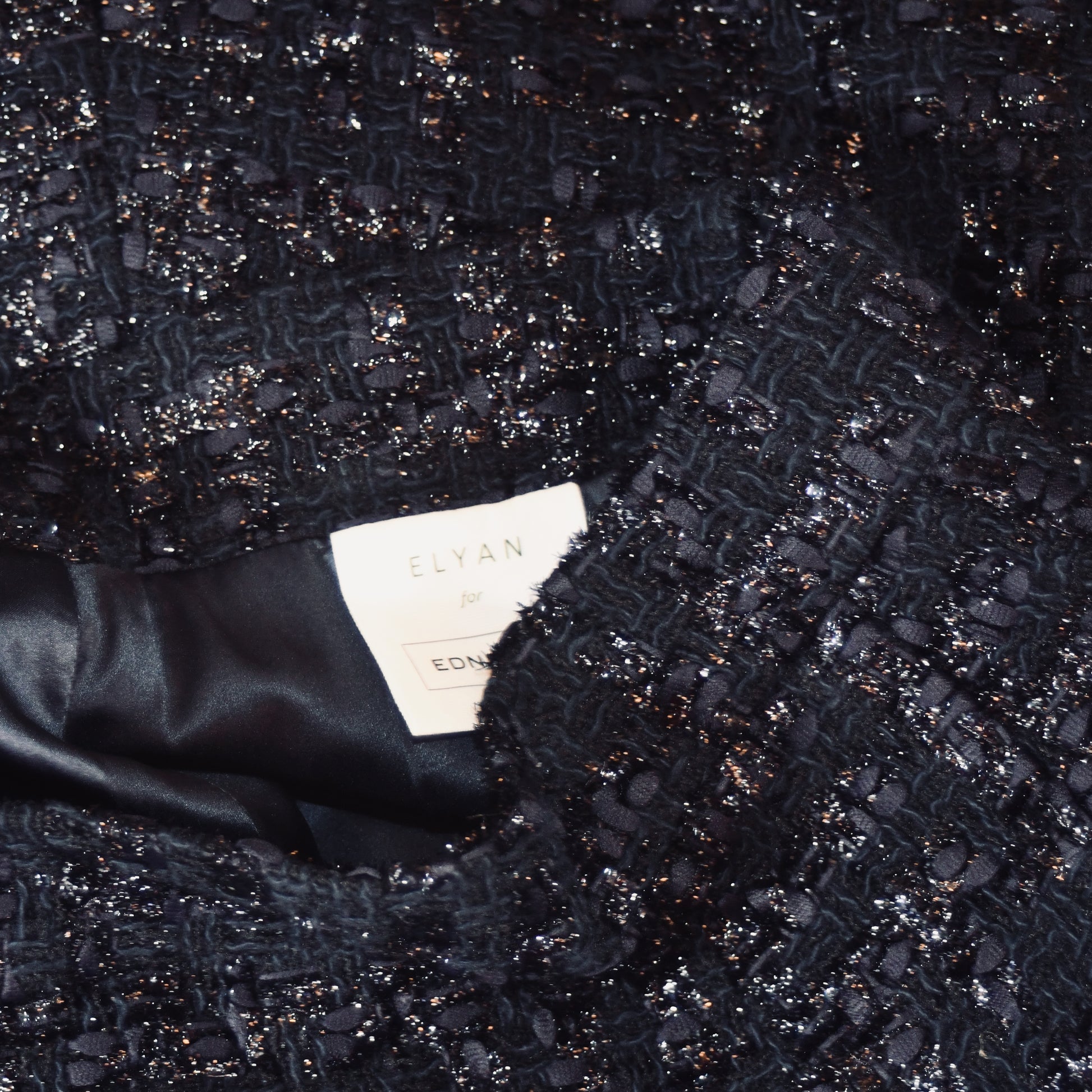Close-up of the textured, dark, sparkly bouclé fabric from EdNerat’s The Waisted A-Line Skirt in Boucle. The smooth black lining contrasts with the textured outer material. Visible clothing label reads ELYAN for EDNA.