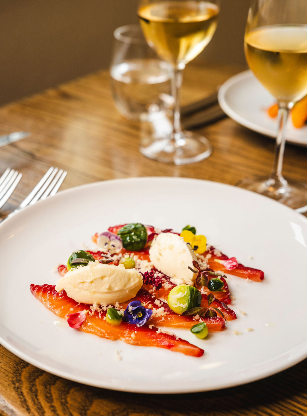 A white plate featuring an elegant, British-made dessert with thinly sliced strawberries, two quenelles of cream, edible flowers, green garnish, and crumb topping. Wine and water glasses are in the background on a wooden table.
