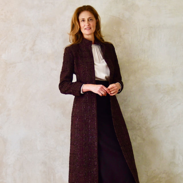 A woman stands against a textured beige wall, wearing a tailored dark coat over a white blouse and black skirt. She has long, wavy brown hair and smiles gently at the camera, highlighting her refined British-made ensemble.