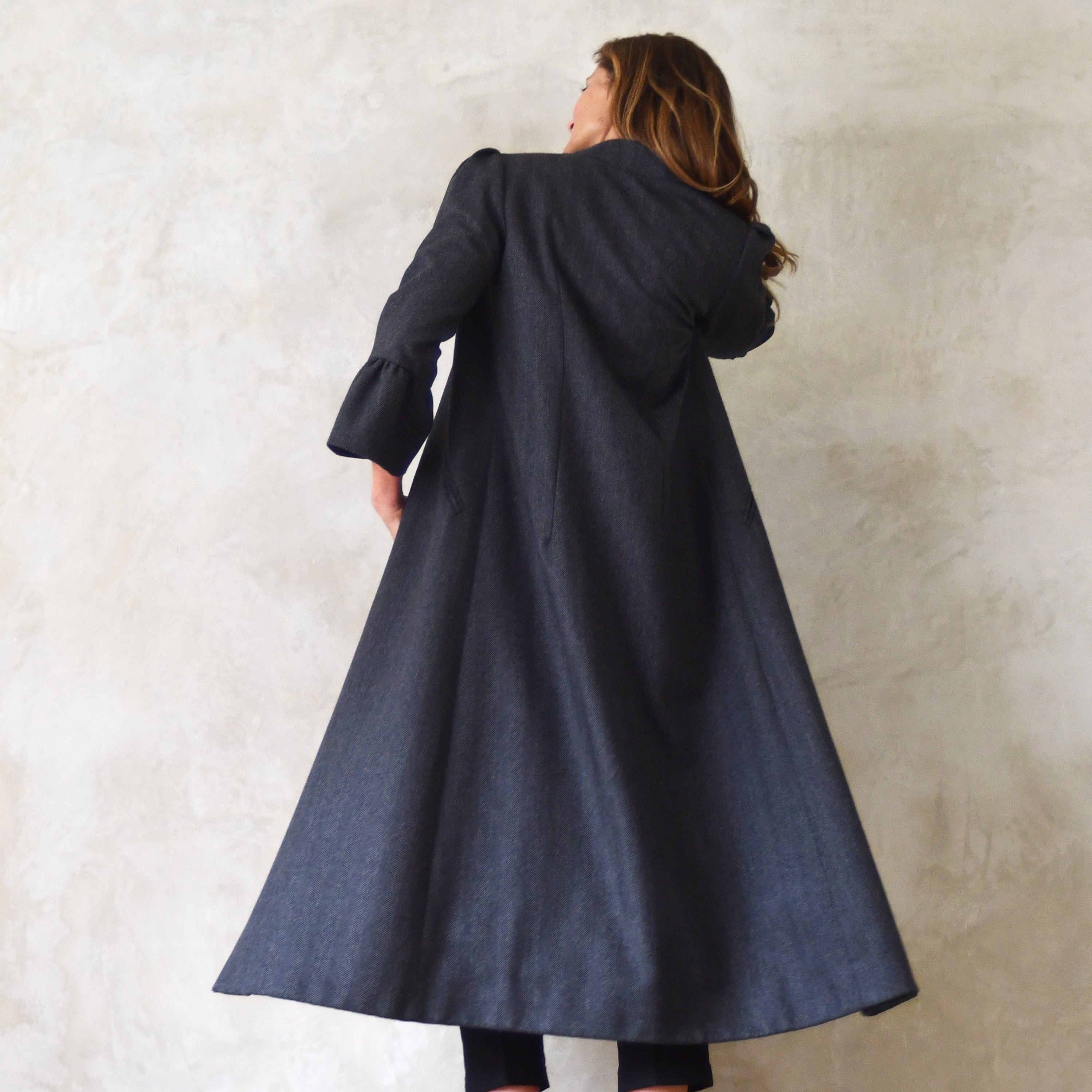 Bell Sleeve A-Line Coat in Charcoal Cashmere Mix Flannel. Bespoke longer length deep grey-coloured A-line coat with gathered shoulder detail, frill cuffs, two slanted pockets to the front and a luscious pink silk lining. Handmade in Britain. Woven in Yorkshire. Rear View.
